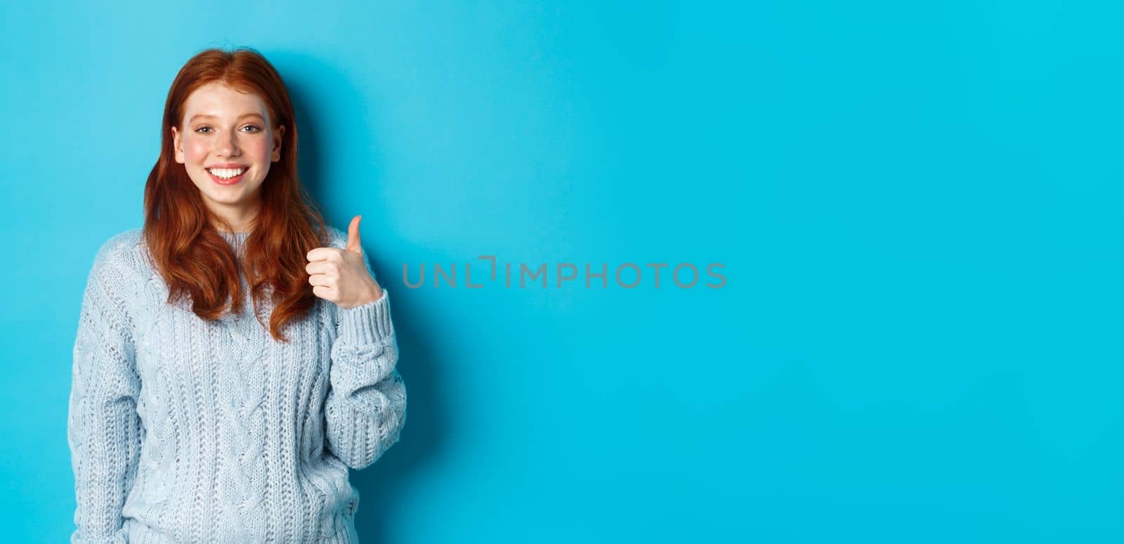 Cute redhead girl in sweater showing thumb up, like and agree, smiling pleased, standing against blue background.