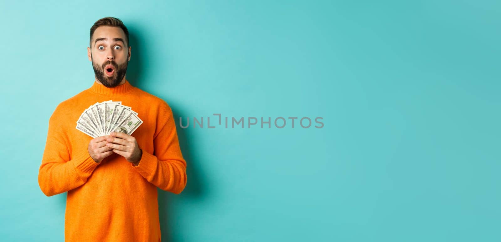 Photo of surprised guy holding money, looking amazed, standing with dollars against turquoise background. Copy space