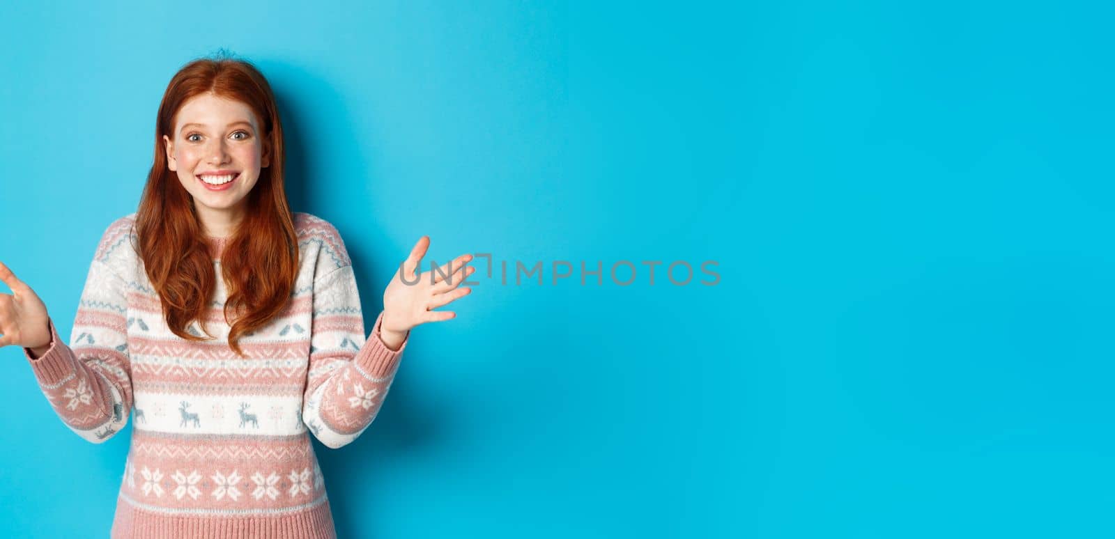 Image of happy redhead girl holding big size object, looking amazed and smiling, receiving Christmas gift, standing over blue background by Benzoix