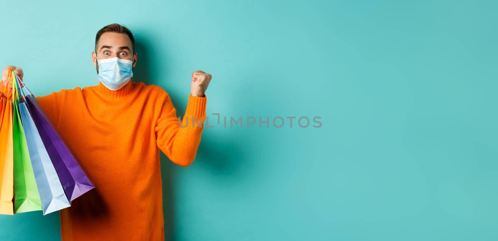 Covid-19, social distancing and lifestyle concept. Excited man in face mask showing shopping bags and rejoicing from discounts, standing over turquoise background by Benzoix