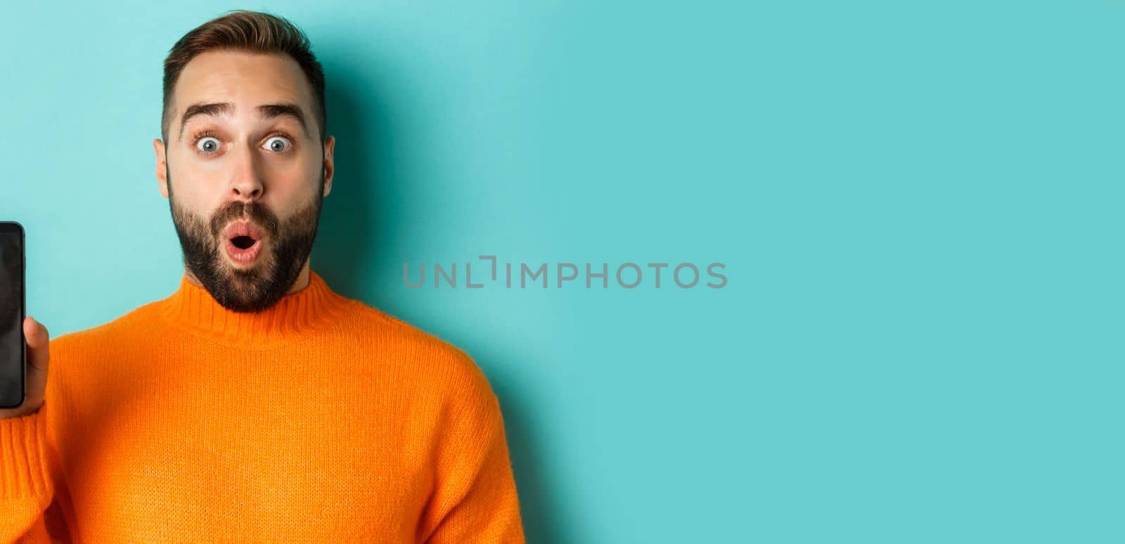 Close-up of handsome bearded guy in orange sweater, showing smartphone screen and smiling, showing promo online, turquoise background by Benzoix