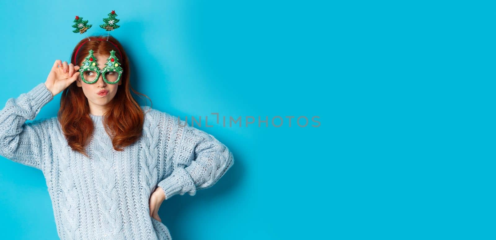 Winter holidays and Christmas sales concept. Beautiful redhead female model celebrating New Year, wearing funny party headband and glasses, smiling silly, blue background.