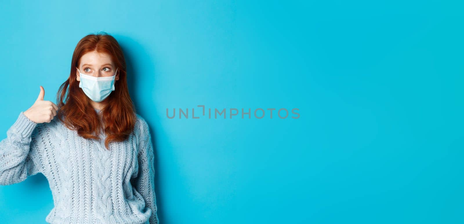 Winter, covid-19 and social distancing concept. Satisfied young redhead woman in face mask showing alright, thumb up gesture and looking left at promo, blue background.