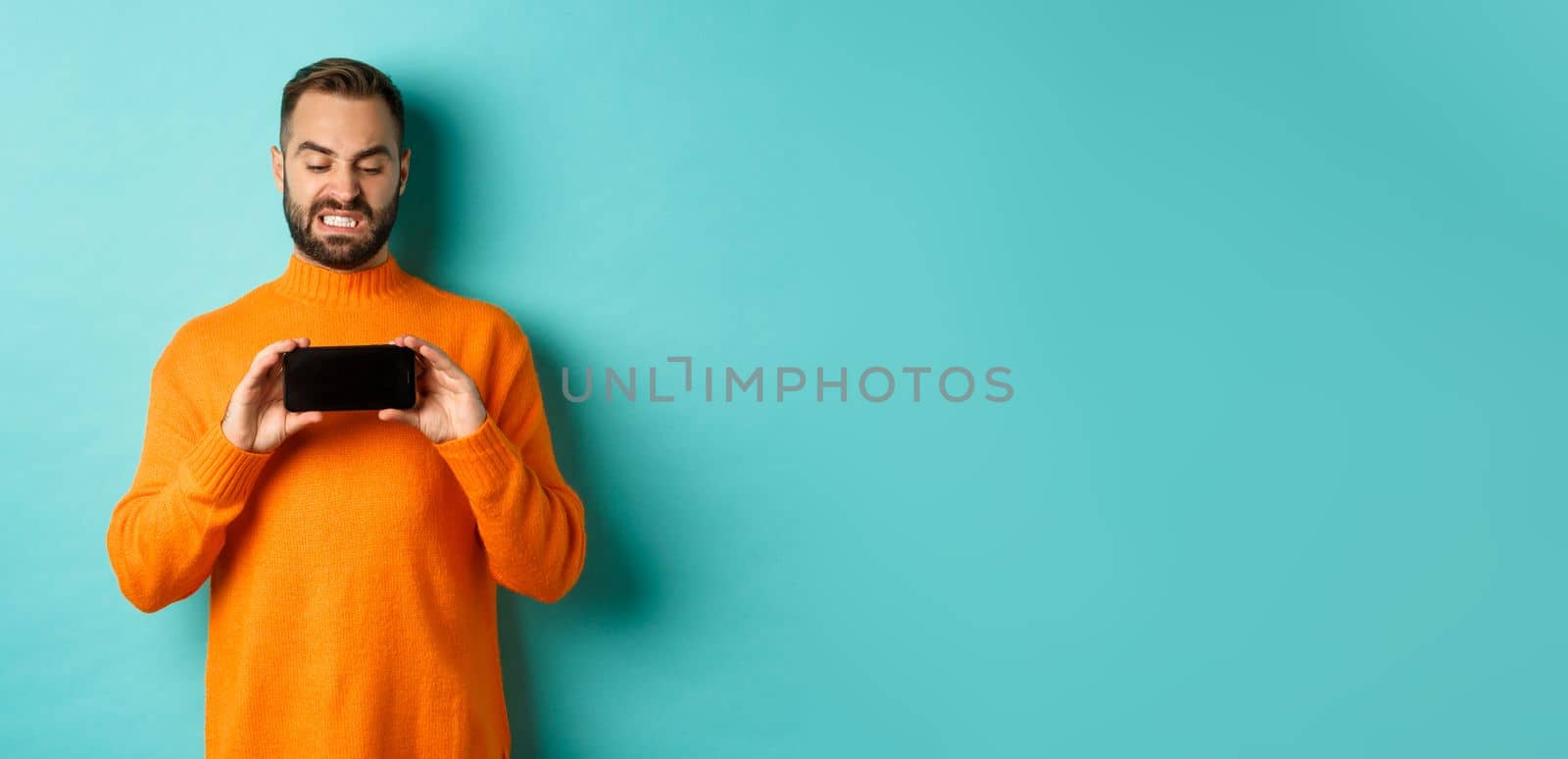 Image of man cringe at something on mobile screen, staring disgusted at display, showing smartphone, standing over light blue background by Benzoix