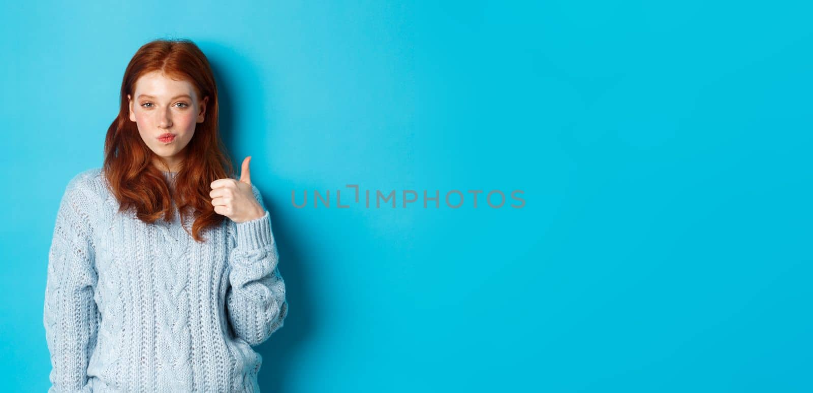 Sassy redhead girl in sweater, looking pleased and showing thumb up, like and agree, standing over blue background.