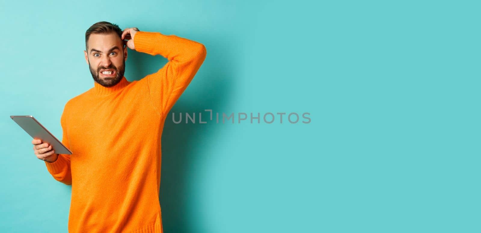 Confused man scratching head and looking worried, working on digital tablet, standing against turquoise background.