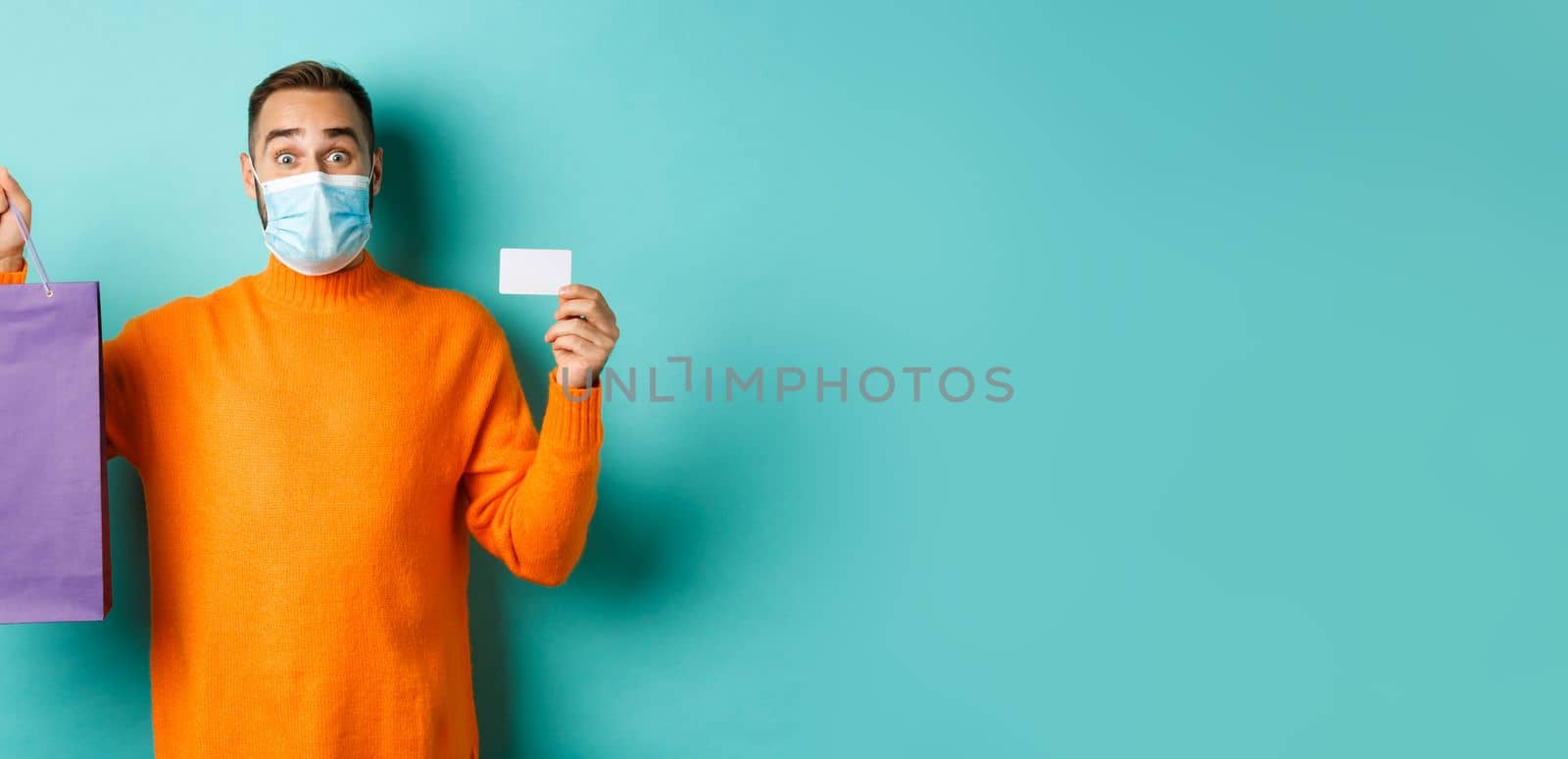 Covid-19, pandemic and lifestyle concept. Happy male customer showing credit card and purple shopping bag, standing over light blue background.