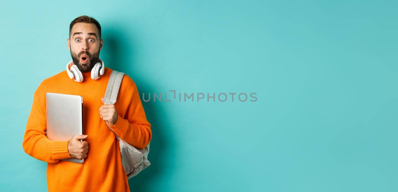 Happy man with backpack and headphones, holding laptop and smiling, looking surprised, standing over turquoise background.