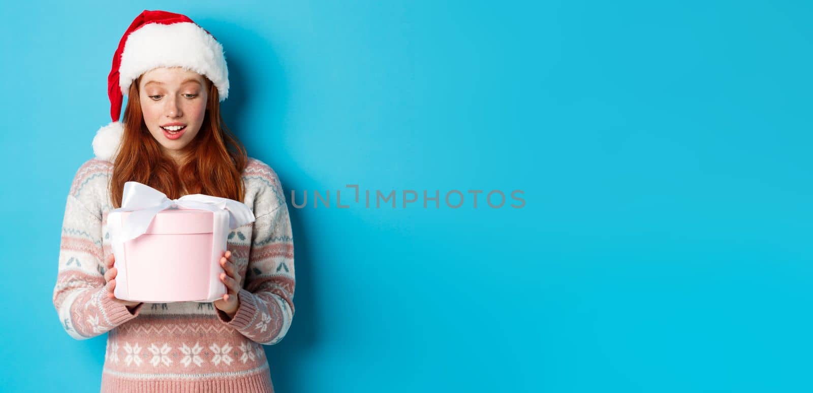 Winter and Christmas Eve concept. Touched and flattered redhead girl looking at box with xmas gift, smiling amazed, standing in santa hat against blue background by Benzoix