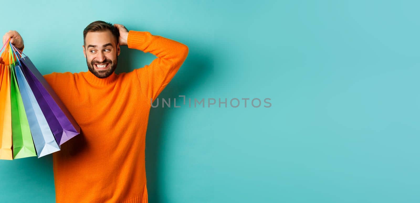 Handsome young man looking awkward and scratching head, staring at shopping bags with gifts, standing over turquoise background by Benzoix