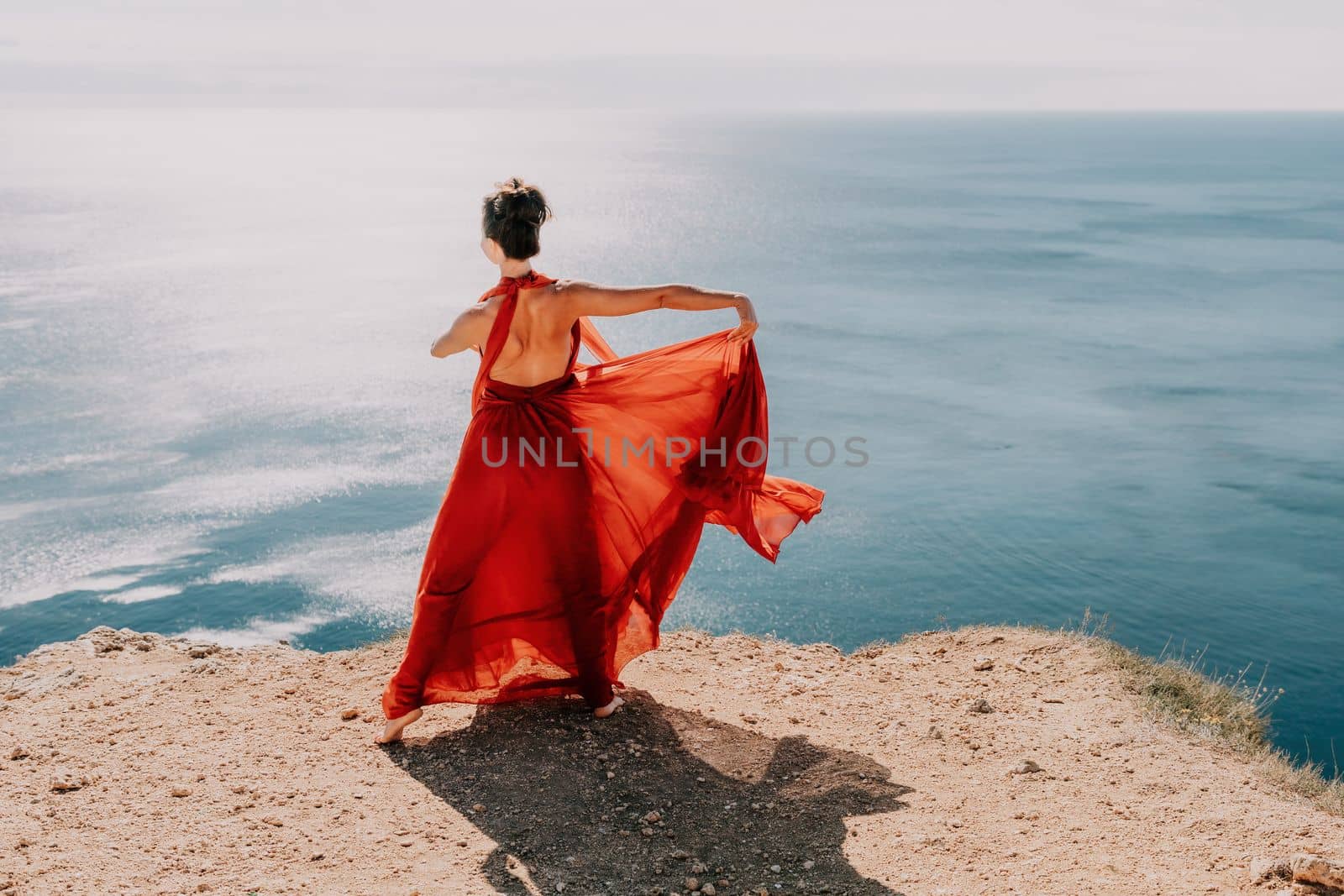 Woman in red dress on sea. Side view a Young beautiful sensual woman in a red long dress posing on a rock high above the sea on sunset. Girl on the nature on blue sky background. Fashion photo. by panophotograph