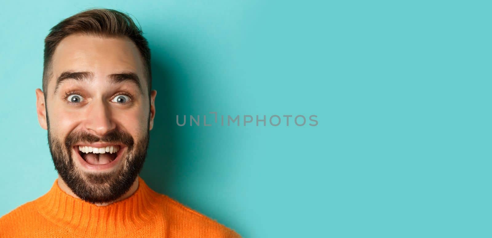 Headshot of handsome caucasian man with beard smiling happy at camera, staring amazed, standing in orange sweater against turquoise background.