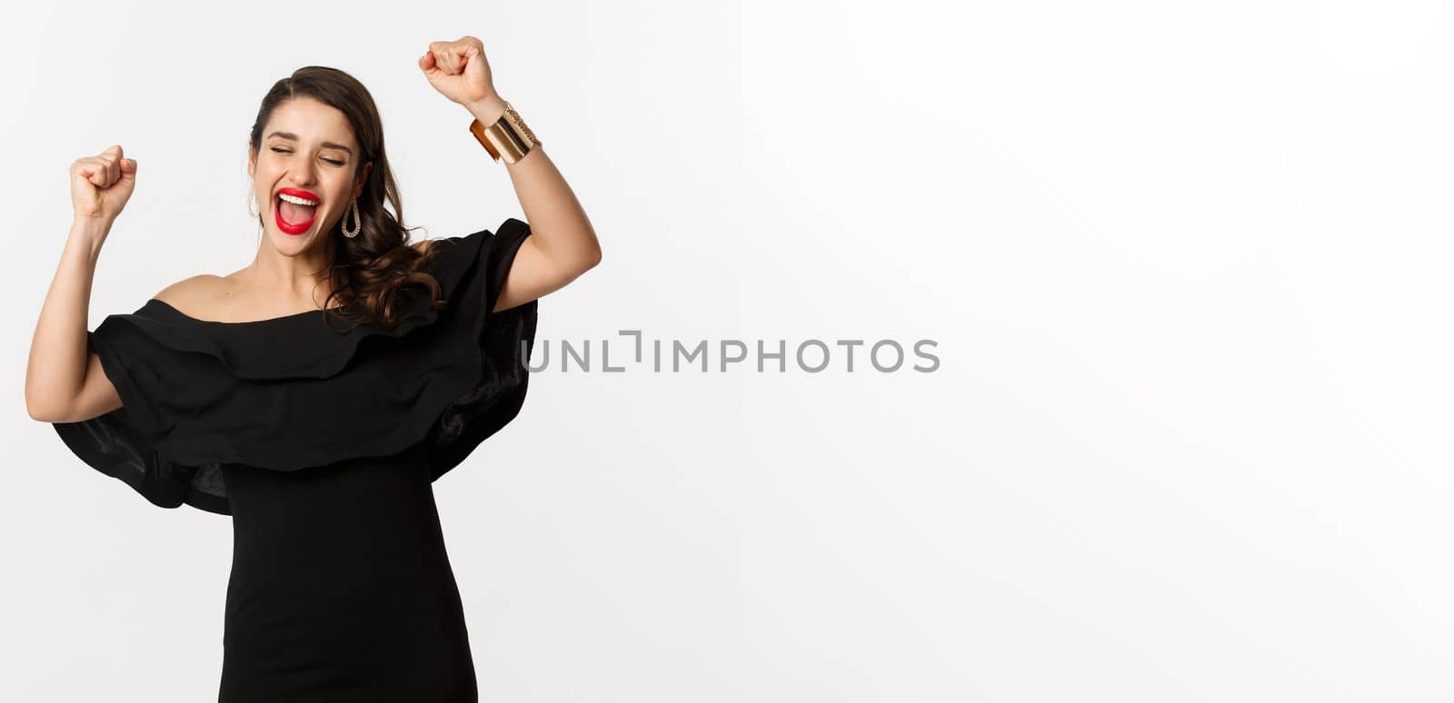 Fashion and beauty. Successful pretty woman in black dress celebrating, rejoicing of winning, triumphing over white background by Benzoix