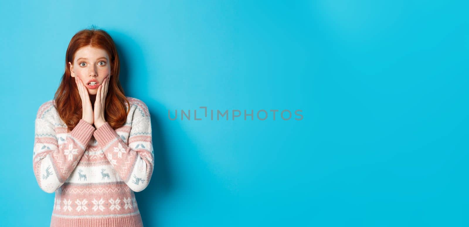 Image of impressed redhead girl gasping and staring at camera, hear big news, standing in winter sweater against blue background.