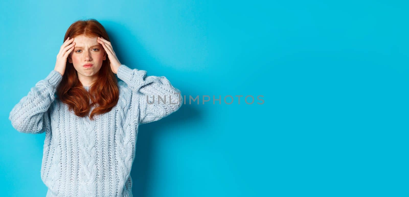 Distressed teenage redhead girl touching head, looking with troubled face expression, standing against blue background, have problem.