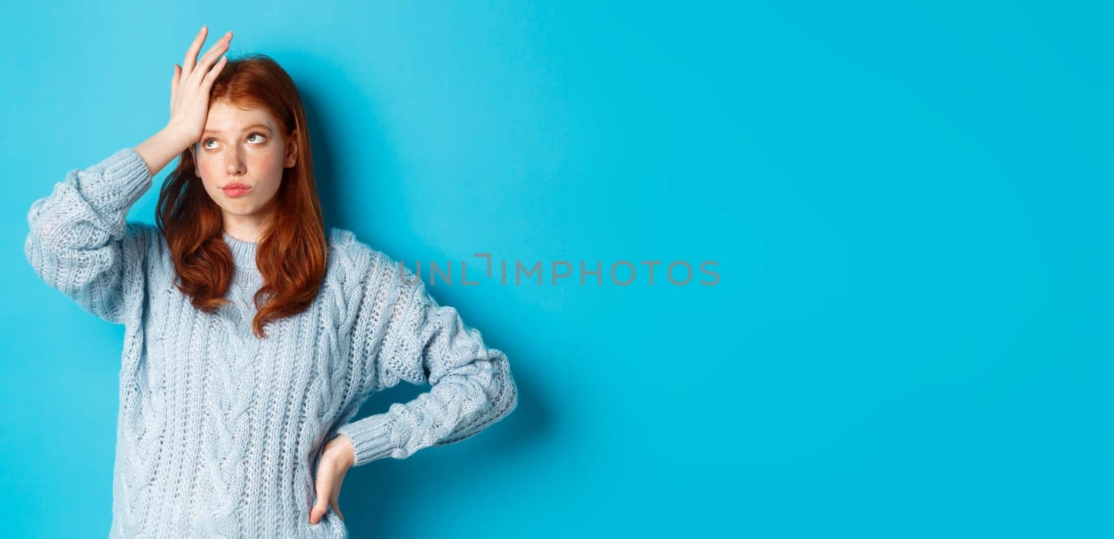 Annoyed and tired redhead teenage girl roll eyes, facepalm and sighing bothered, standing in sweater against blue background by Benzoix