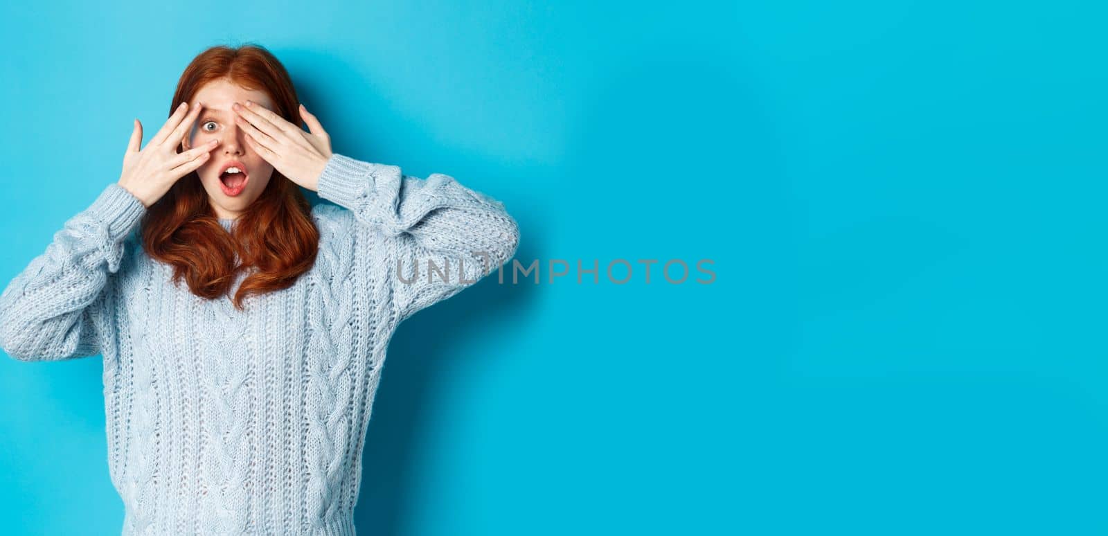 Excited teenage redhead girl open eyes to see holiday surprise, receiving presents, looking amazed at camera, standing over blue background by Benzoix