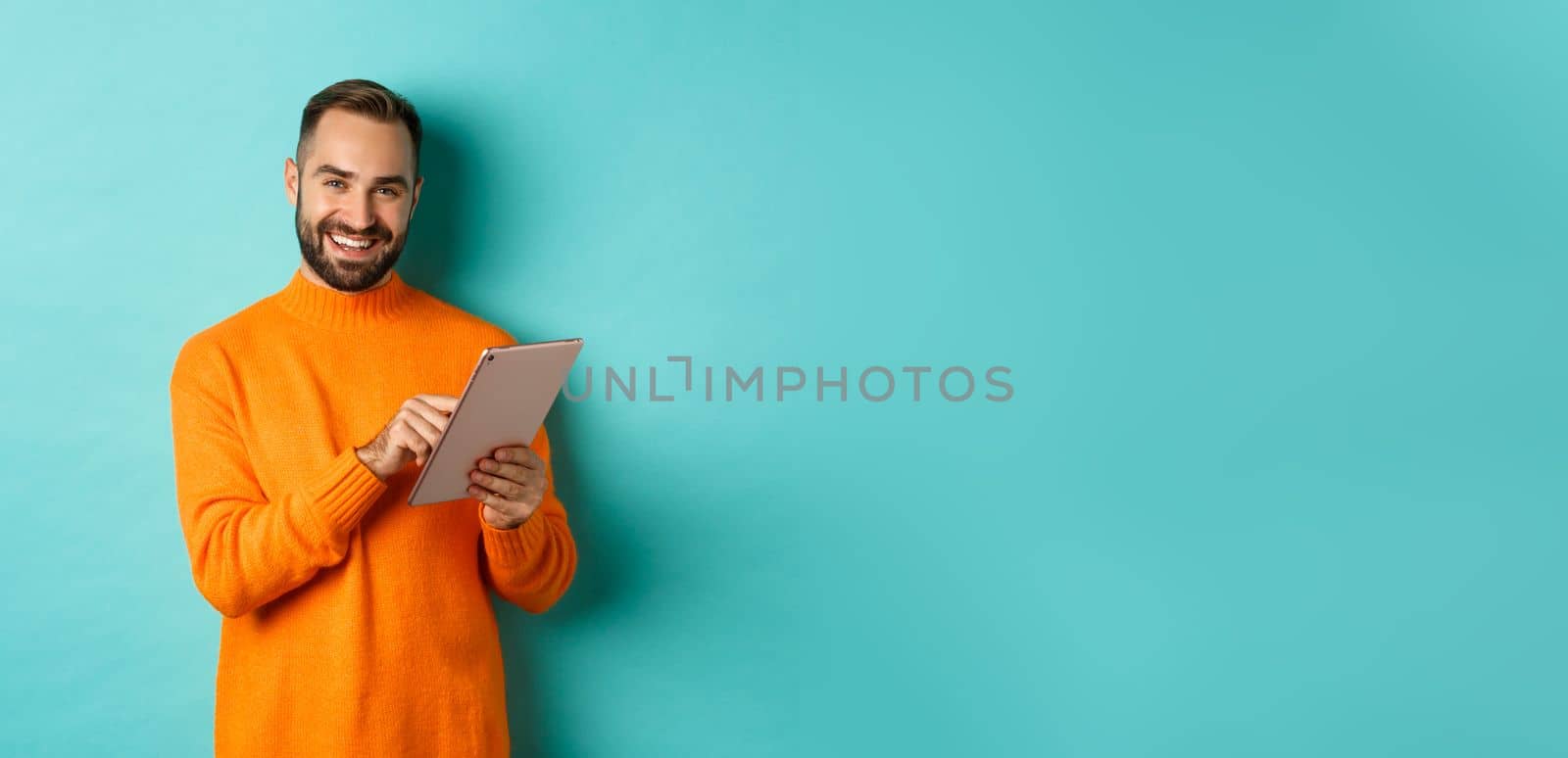 Handsome adult man using digital tablet and smiling, shopping online, standing over light blue background.