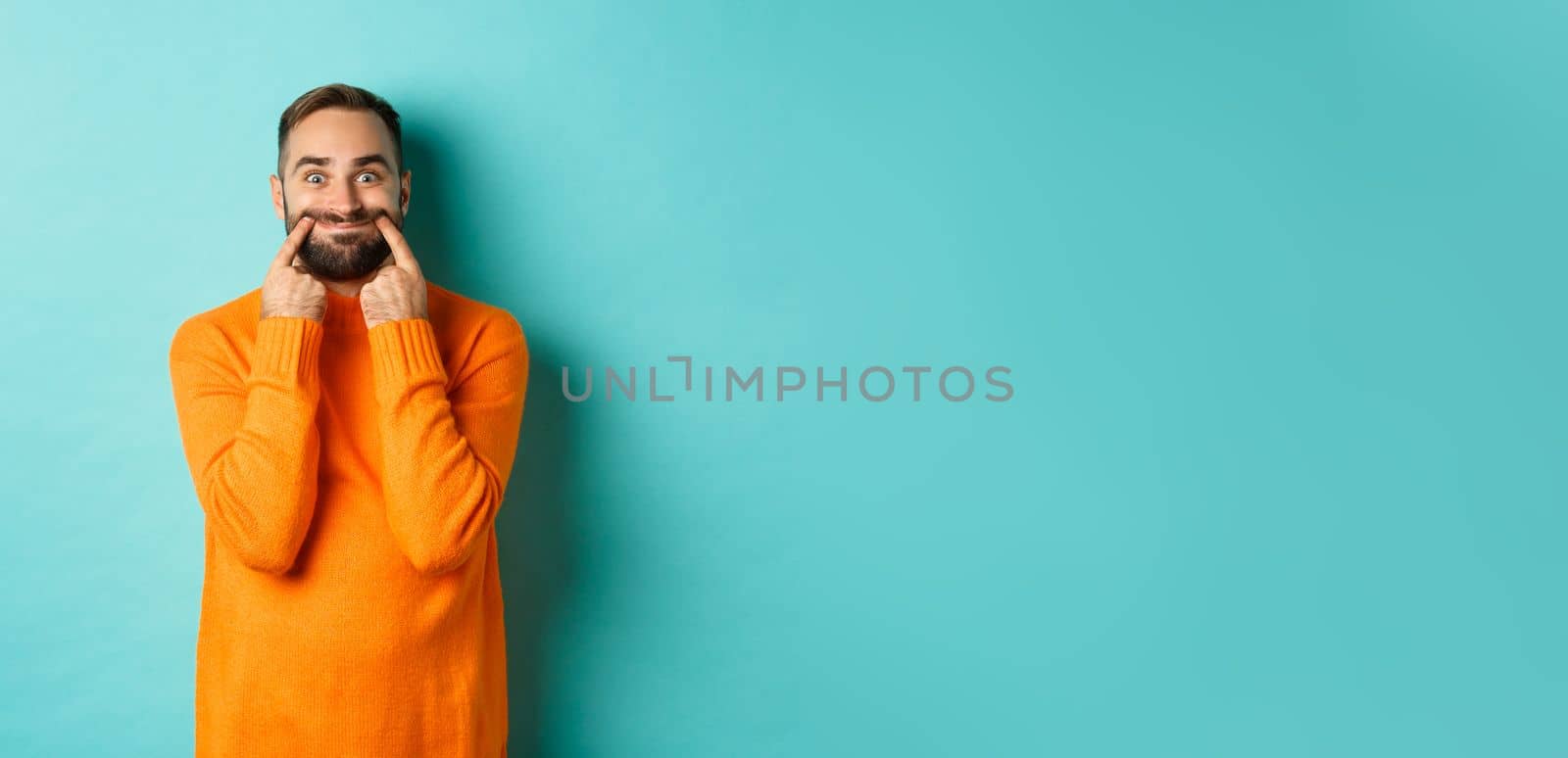 Image of bearded man stretching lips in happy smile, faking happiness, standing over light blue background.