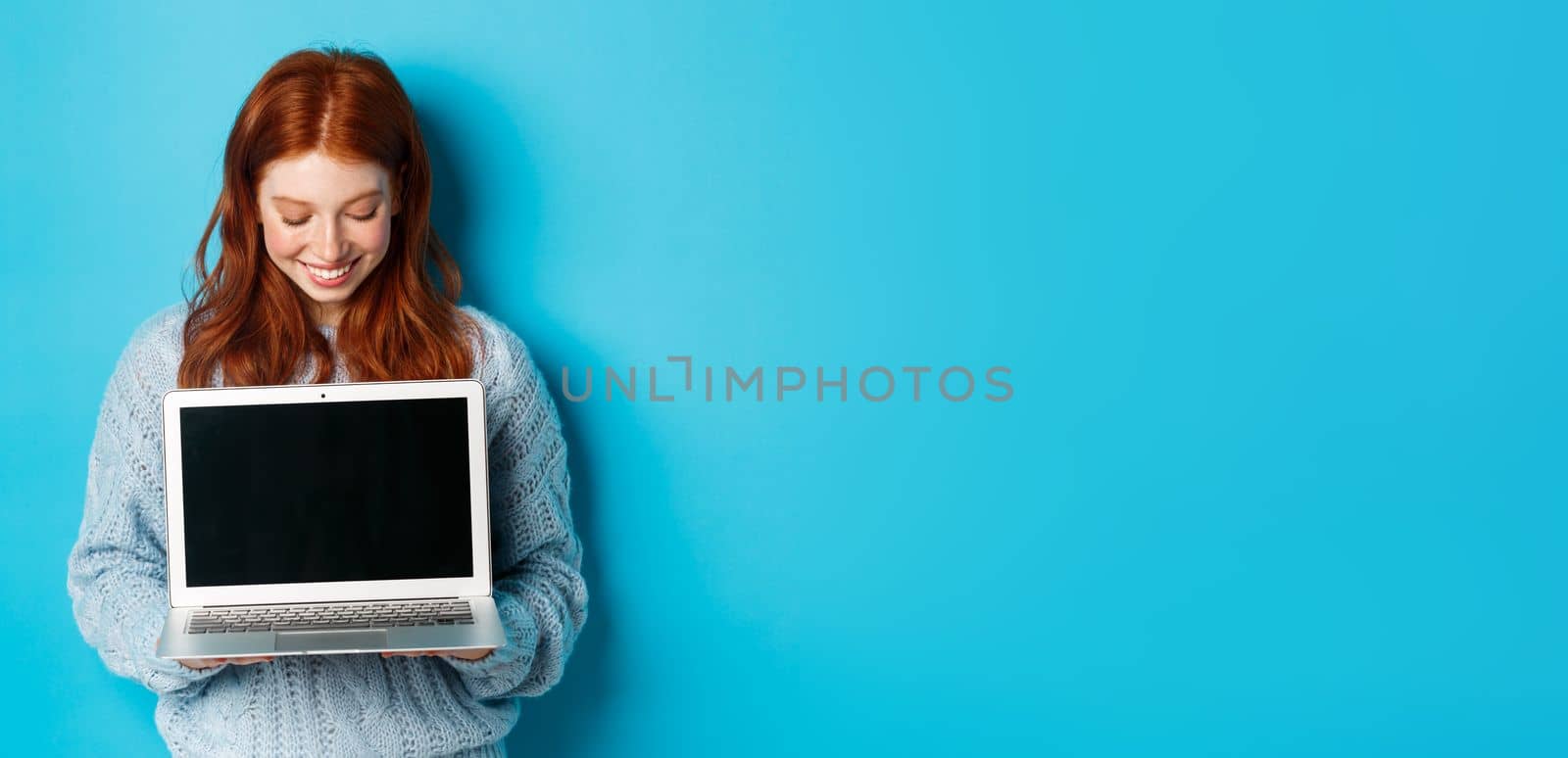 Cute redhead woman in sweater, showing and looking at laptop screen with pleased smile, demonstrating something online, standing over blue background.