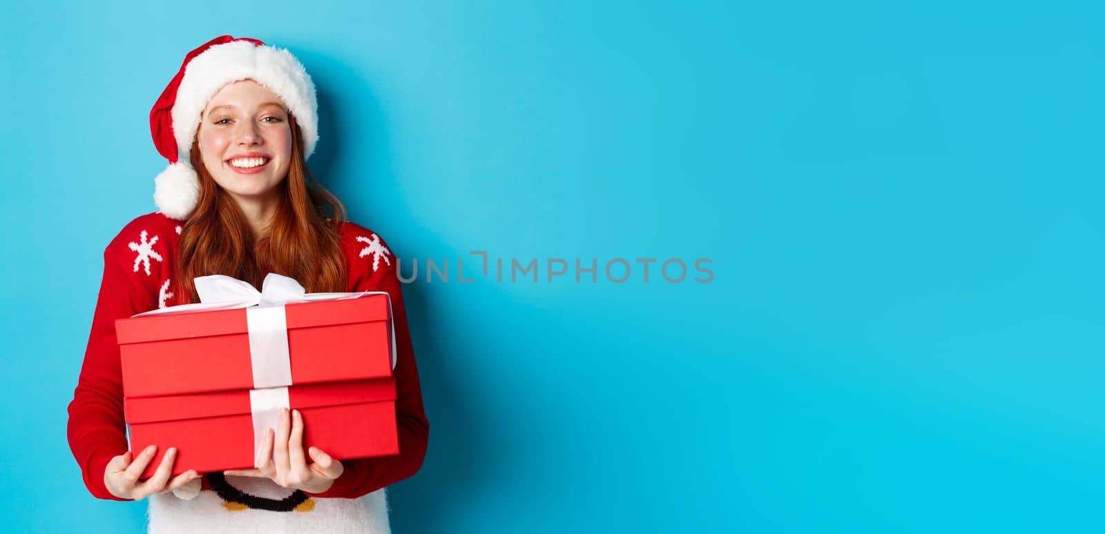 Happy holidays and Christmas concept. Cheerful teen girl with red hair, wearing santa hat and holding xmas presents.