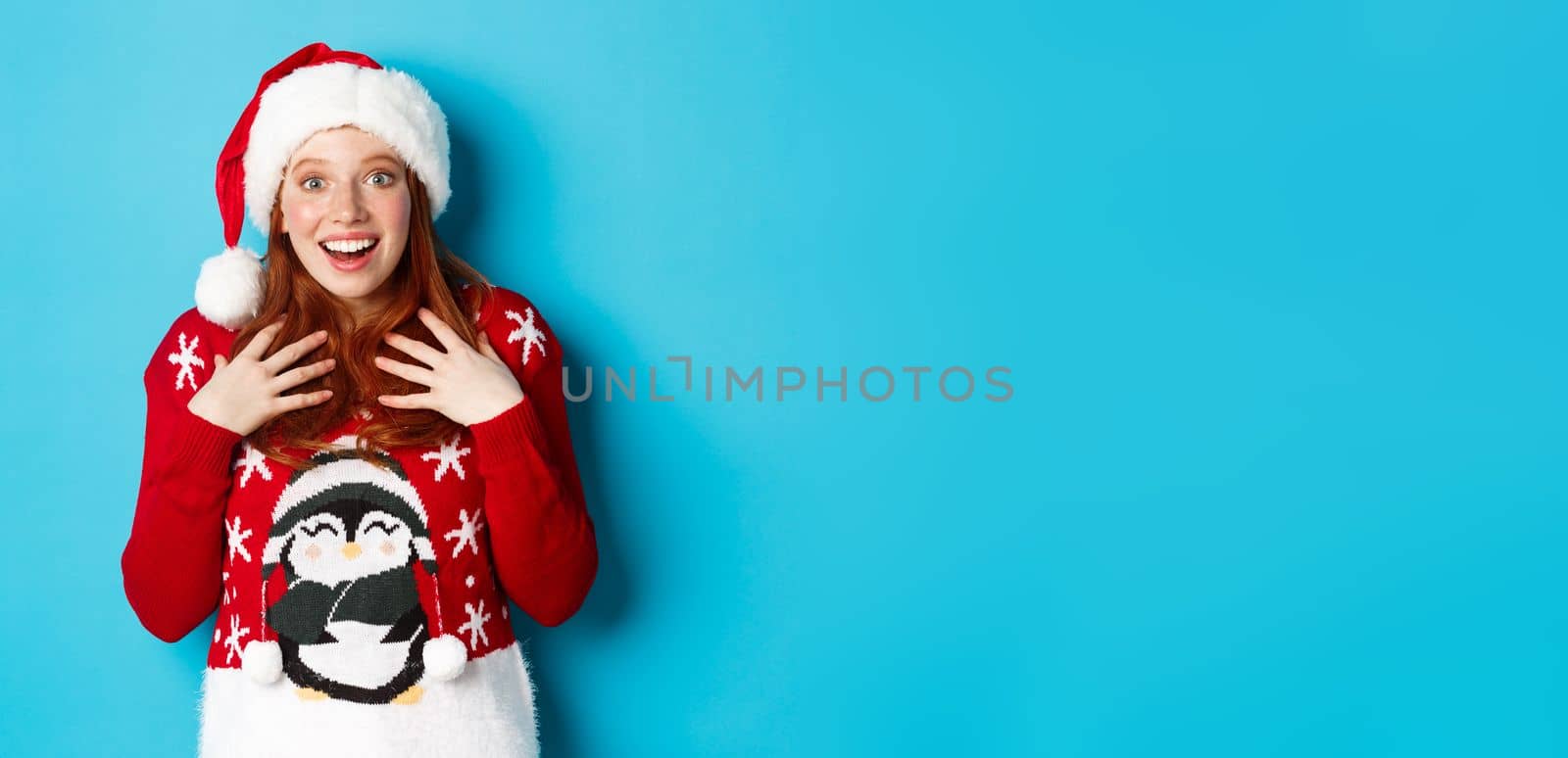 Happy holidays and Christmas concept. Surprised redhead girl receiving unexpected gift, gasping amazed and staring with plesant disbelief, standing in santa hat against blue background by Benzoix