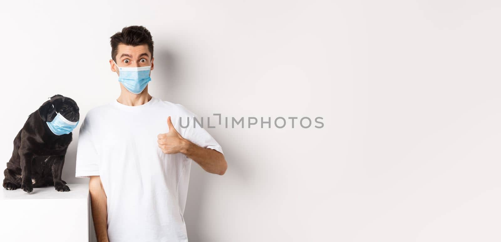 Covid-19, animals and quarantine concept. Image of funny young man and small dog in medical masks, owner showing thumb up in approval, praise something, white background.