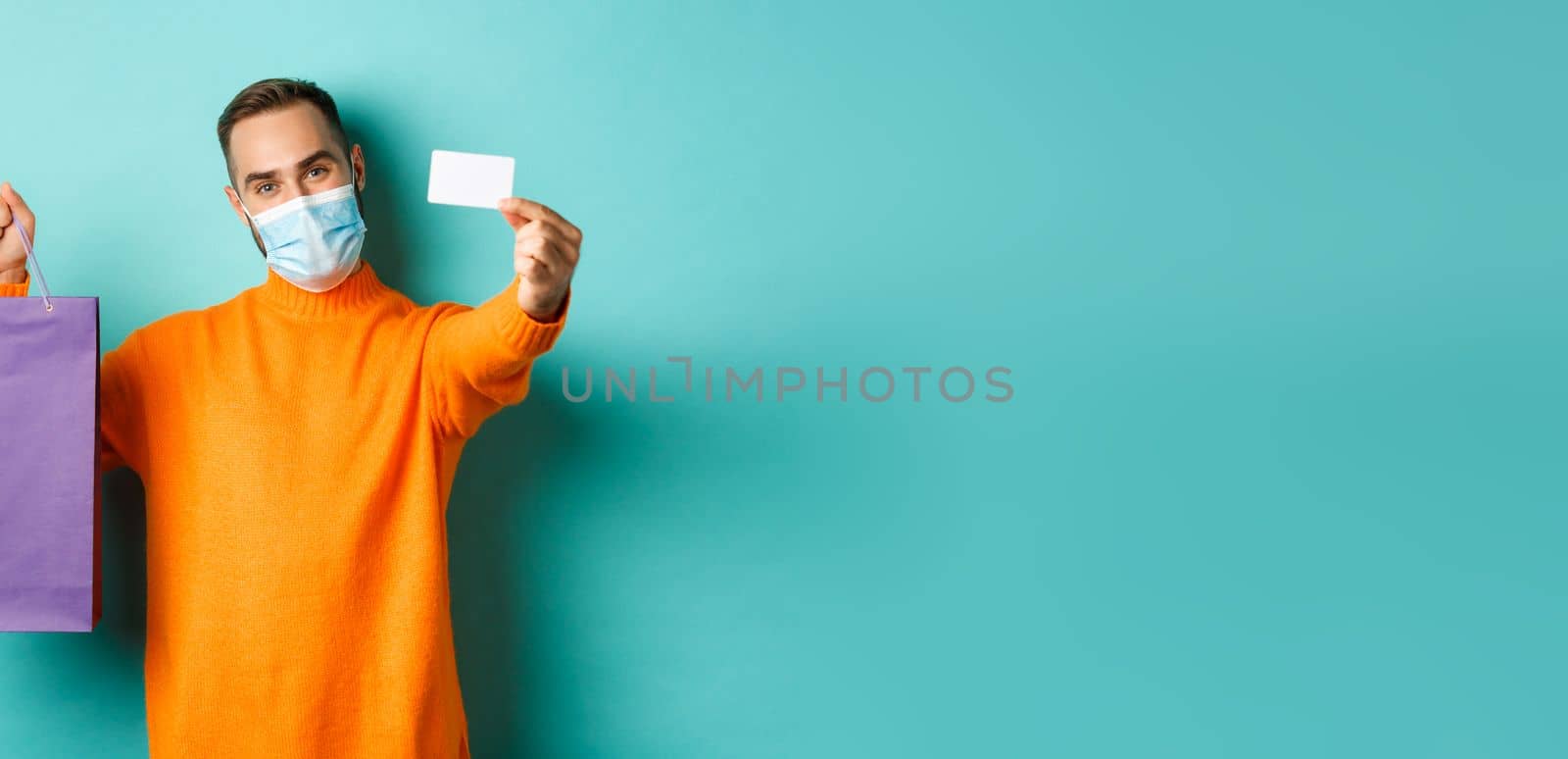 Covid-19, pandemic and lifestyle concept. Happy male customer in face mask showing credit card and purple shopping bag, standing over light blue background.