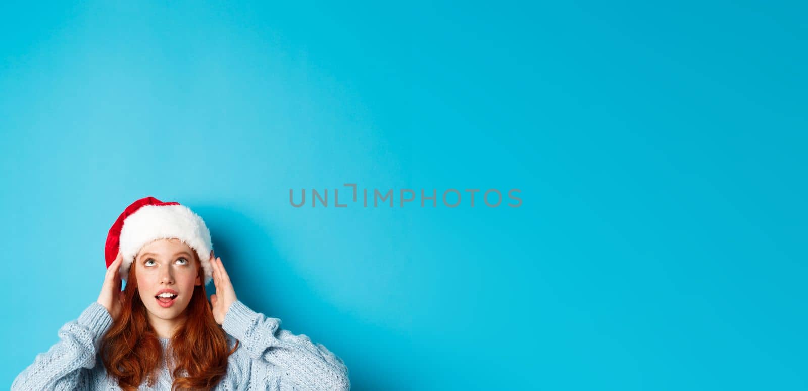 Winter holidays and Christmas eve concept. Head of cute redhead girl in santa hat, appear from bottom and looking up at copy space, staring logo, standing over blue background by Benzoix