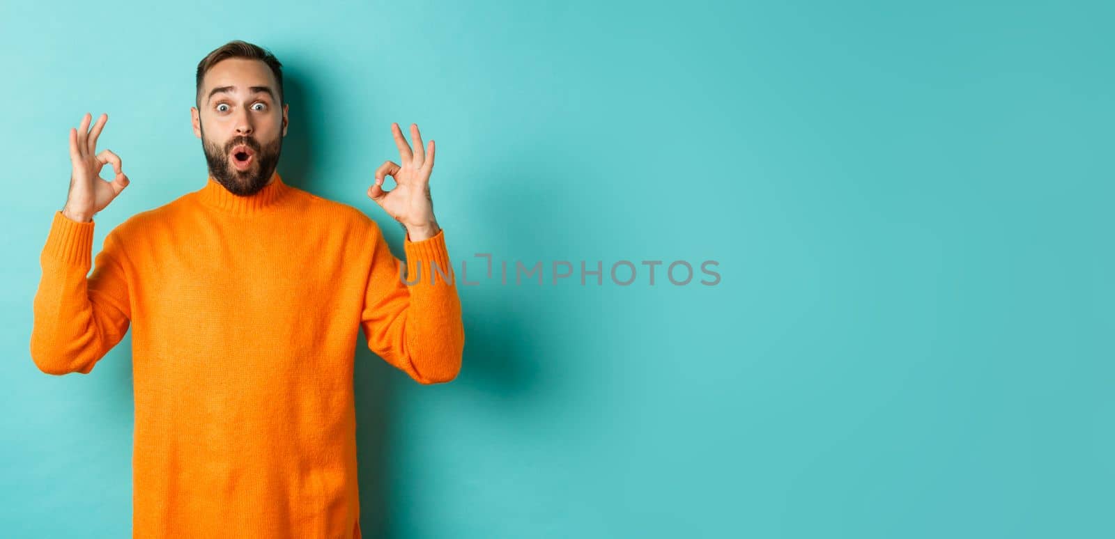 Impressed man praising something super cool, showing okay sign and looking wondered at camera, light blue background.