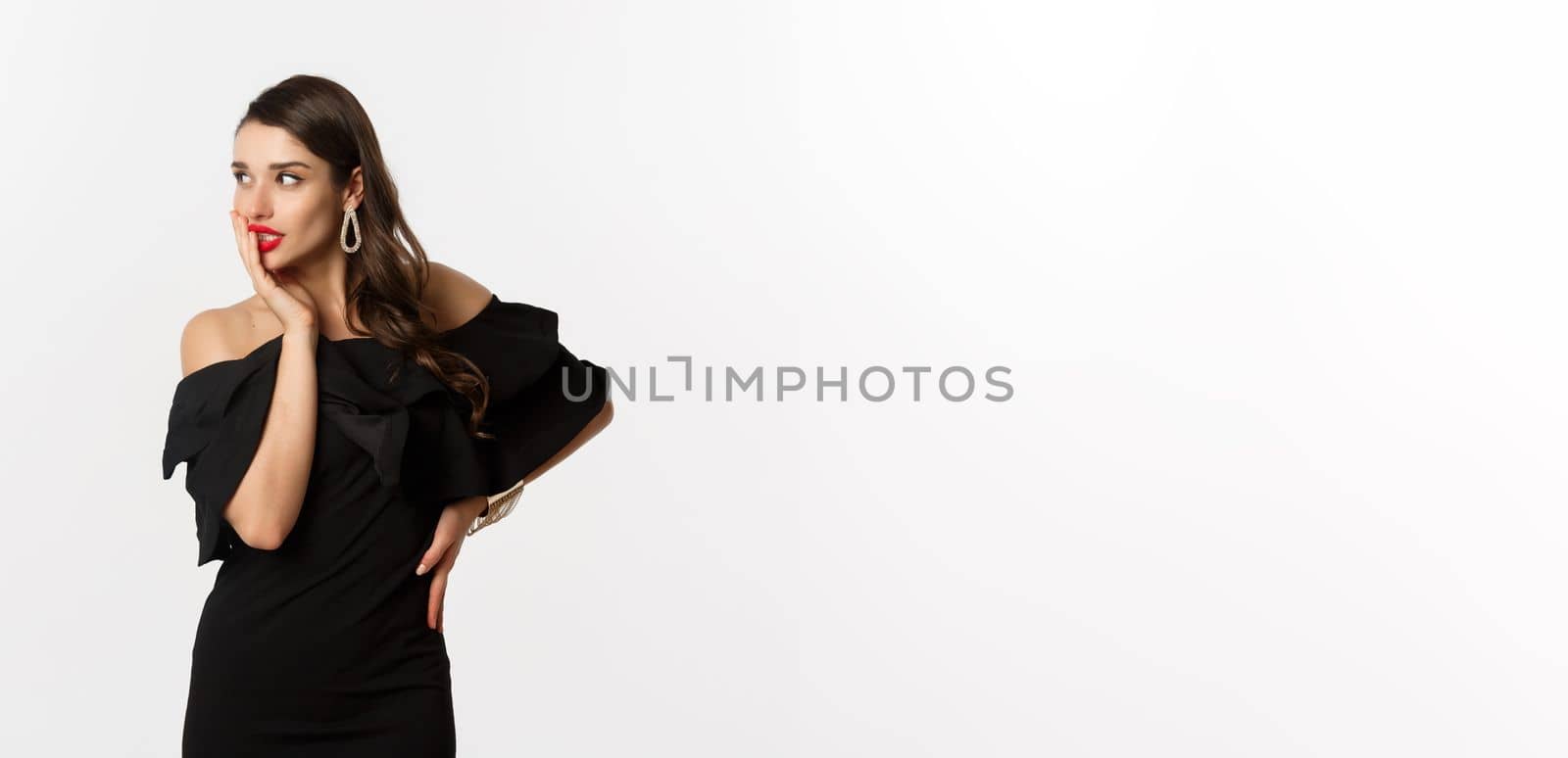 Fashion and beauty. Image of stylish beautiful woman in black dress and makeup, looking left with temptation, touching red lips, standing over white background.