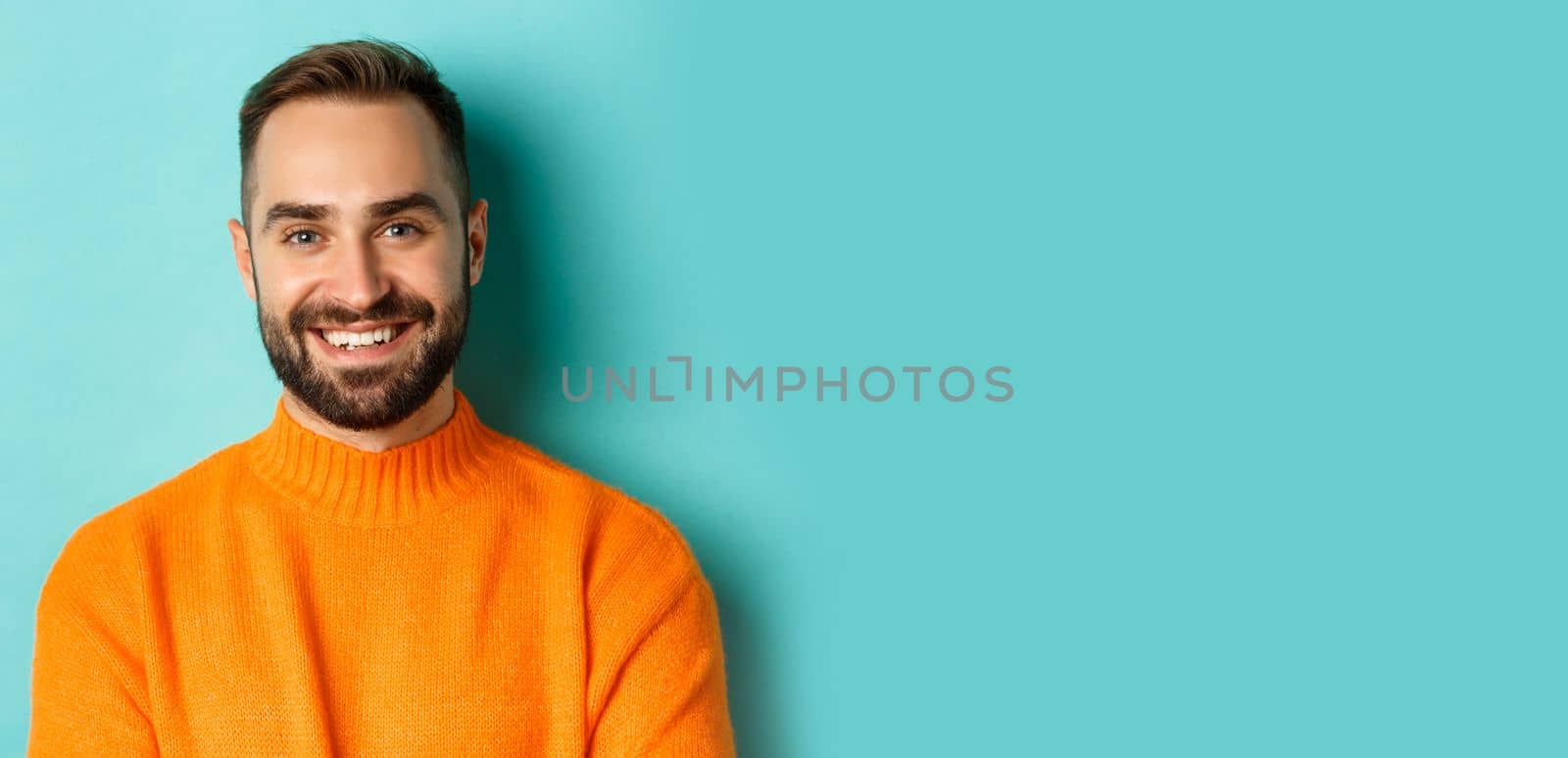 Close-up of handsome caucasian man smiling at camera, looking confident, wearing orange sweater, standing against turquoise background.