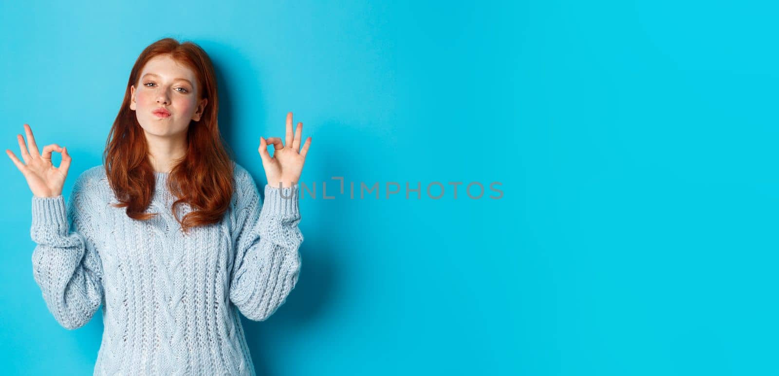 Redhead teenage girl showing okay signs, looking satisfied and proud, agree, give positive answer, praising good choice, standing over blue background by Benzoix
