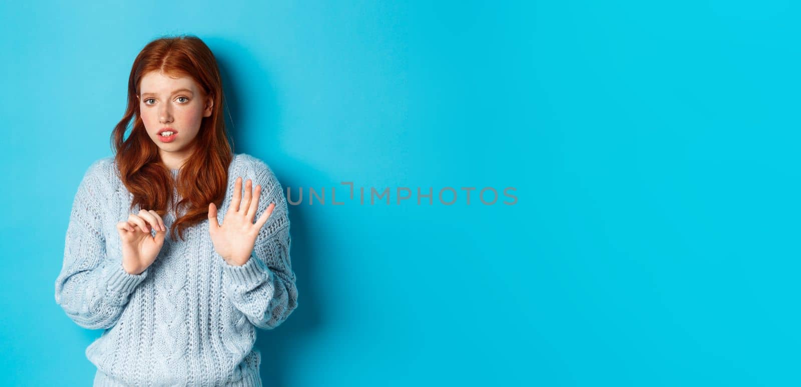Worried redhead girl refusing or declining an offer, shaking hands and looking anxiously at camera, rejecting something unpleasant, standing over blue background.