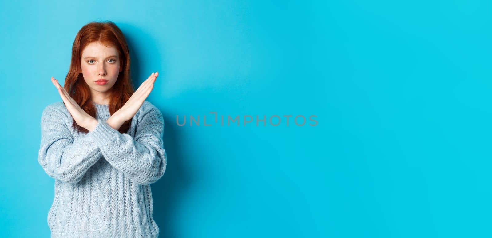 Serious redhead girl looking confident, showing cross gesture to stop and forbid action, standing over blue background by Benzoix