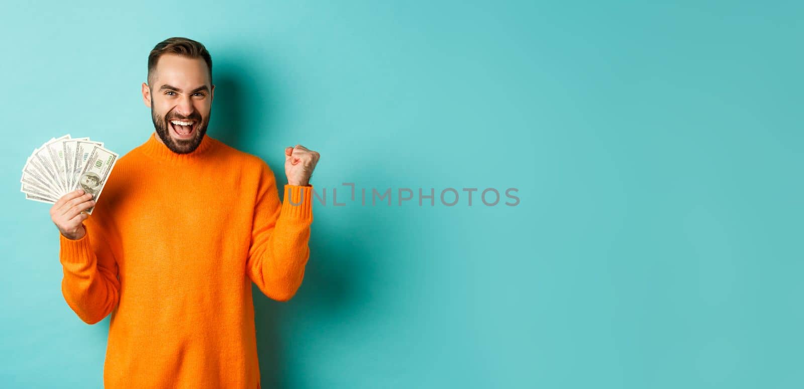 Shopping. Cheerful guy holding money, winning prize in cash and making fist pump, triumphing with satisfied expression, standing in orange sweater against turquoise background by Benzoix