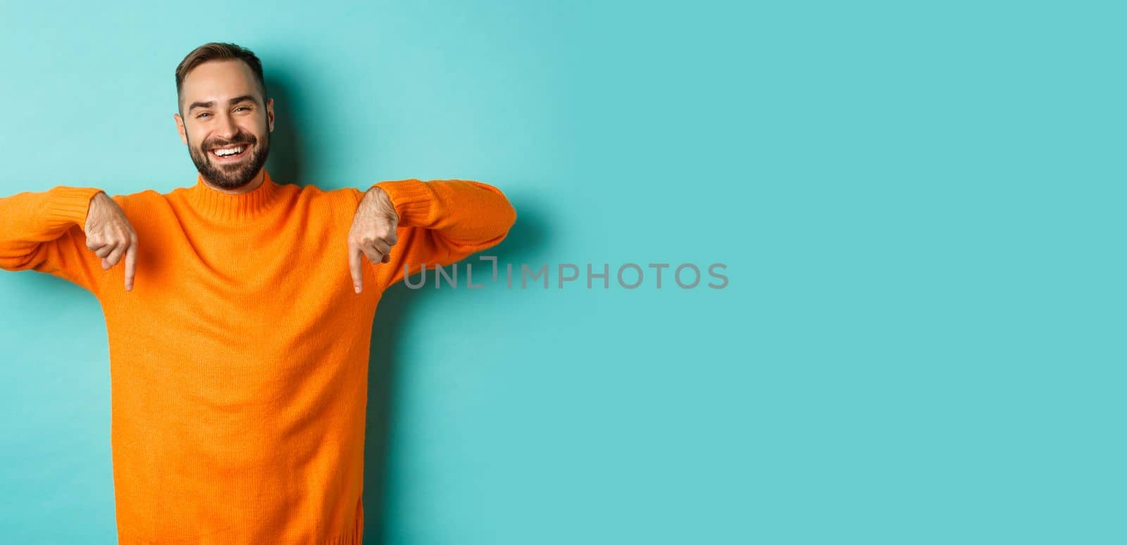 Happy adult man with beard pointing fingers down, smiling cheerful, showing advertisement, standing over turquoise background.