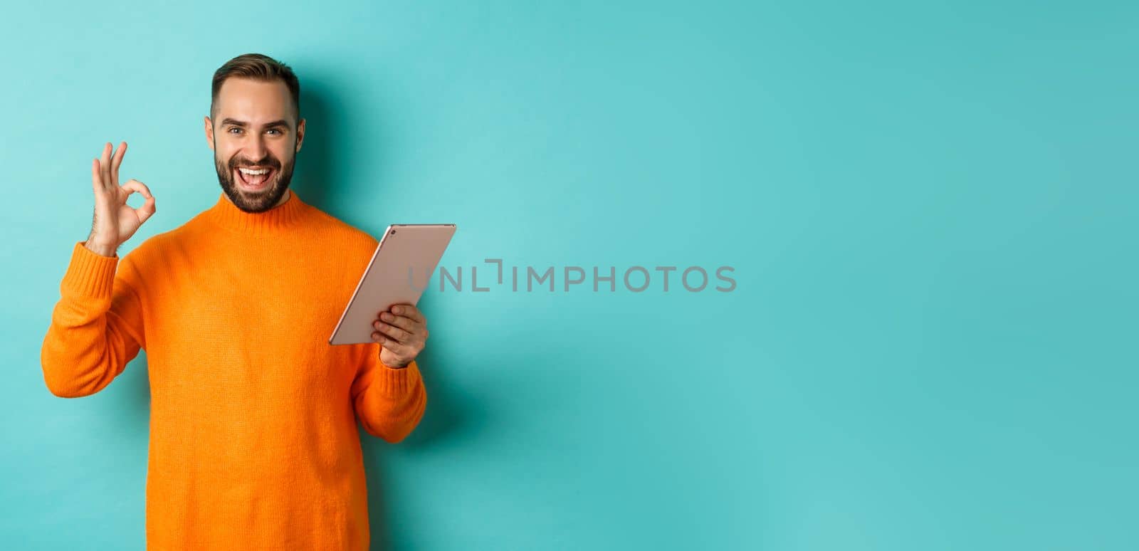 Satisfied adult man smiling, using digital tablet and showing okay sign, approve and agree, standing against turquoise background.