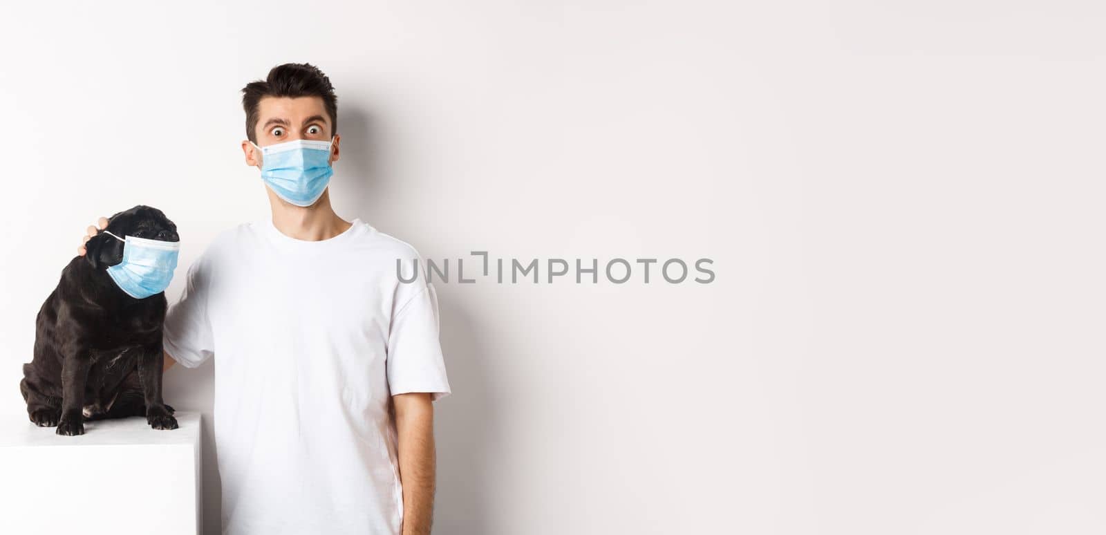 Covid-19, animals and quarantine concept. Image of funny young man and pug in medical masks, staring at camera, standing over white background by Benzoix