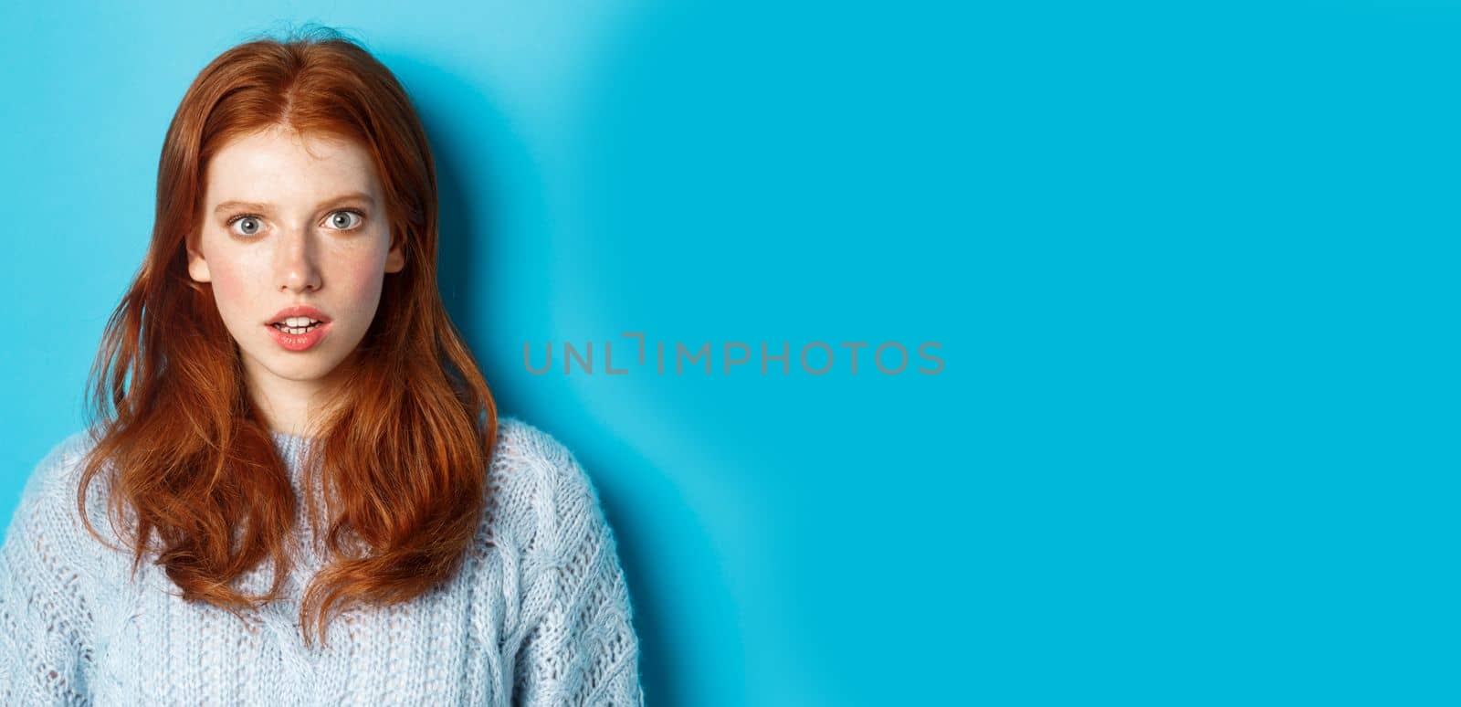 Close-up of amazed redhead girl staring at camera with complete disbelief, standing shocked against blue background.