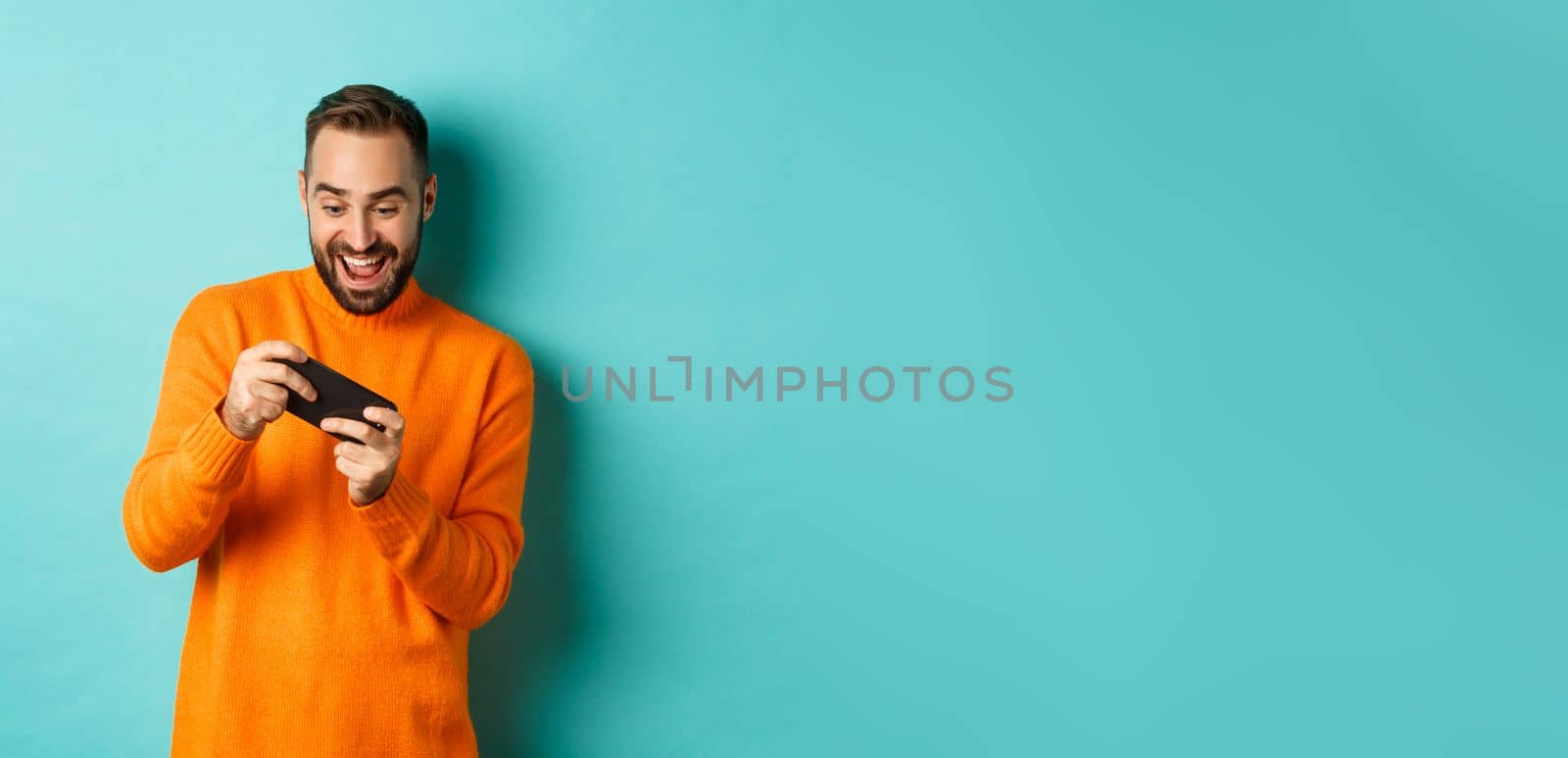 Excited adult man playing on mobile phone, looking amazed at smartphone screen, standing over turquoise background.