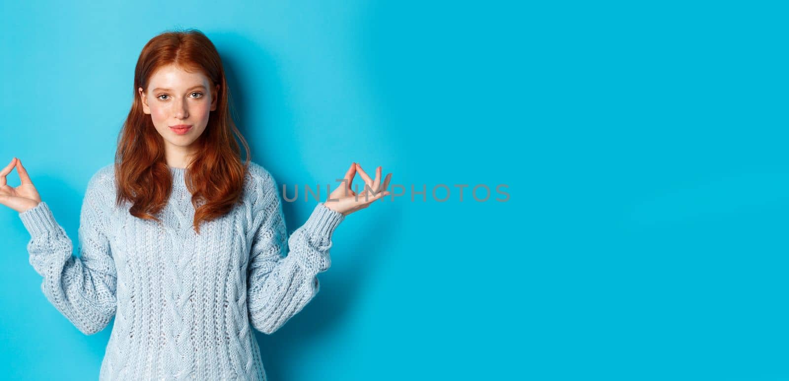 Smiling confident girl with red hair staying patient, holding hands in zen, meditation pose and staring at camera, practice yoga, standing calm against blue background.