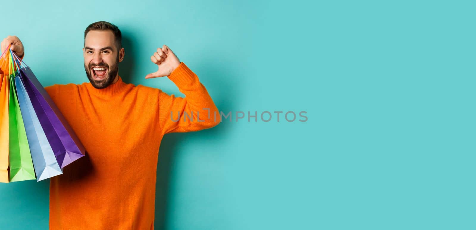 Handsome bearded man in orange sweater, buying gifts, pointing at himself and showing shopping bags, standing over blue background.