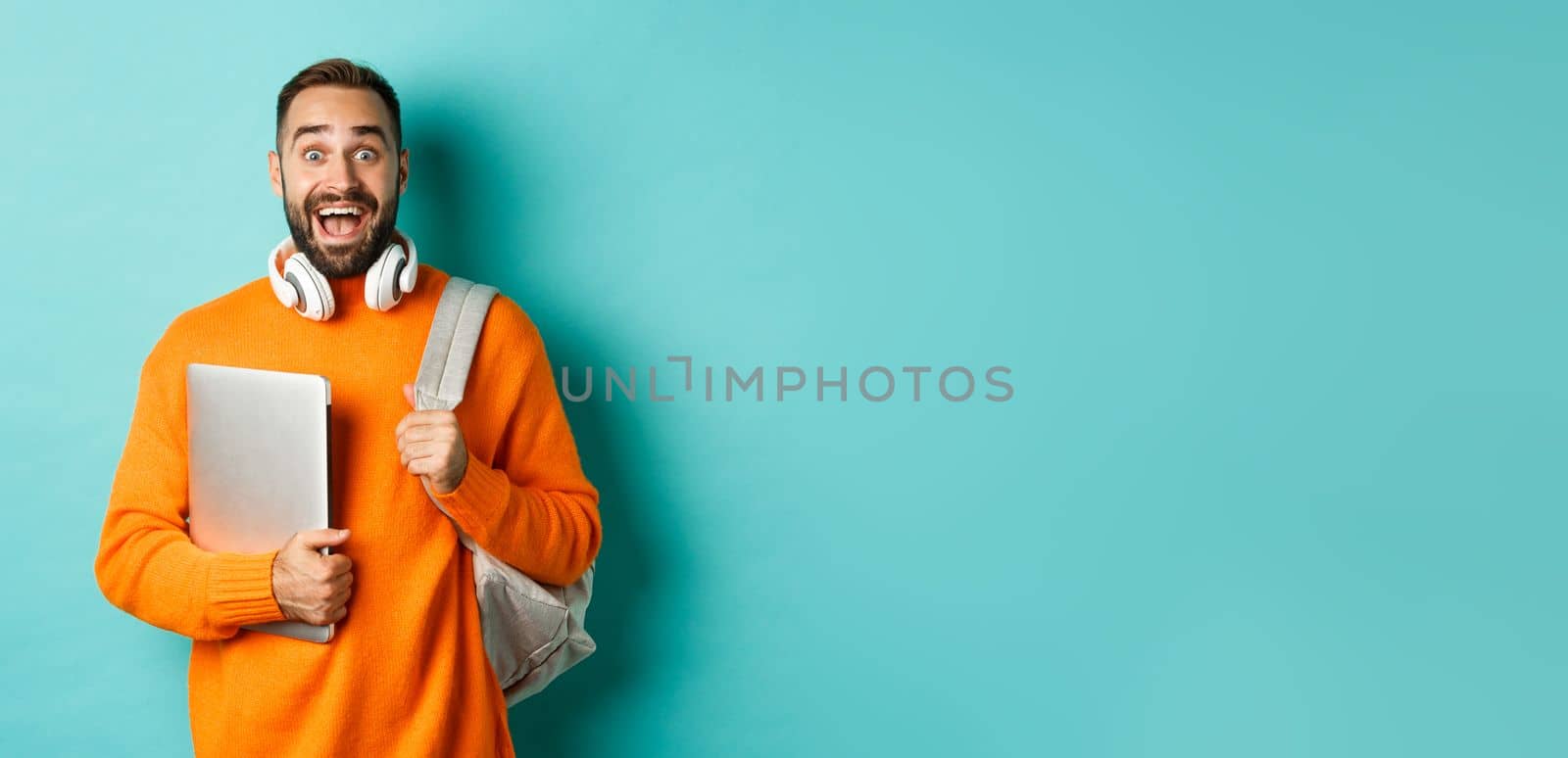 Happy man with backpack and headphones, holding laptop and smiling, looking excited, standing over turquoise background by Benzoix