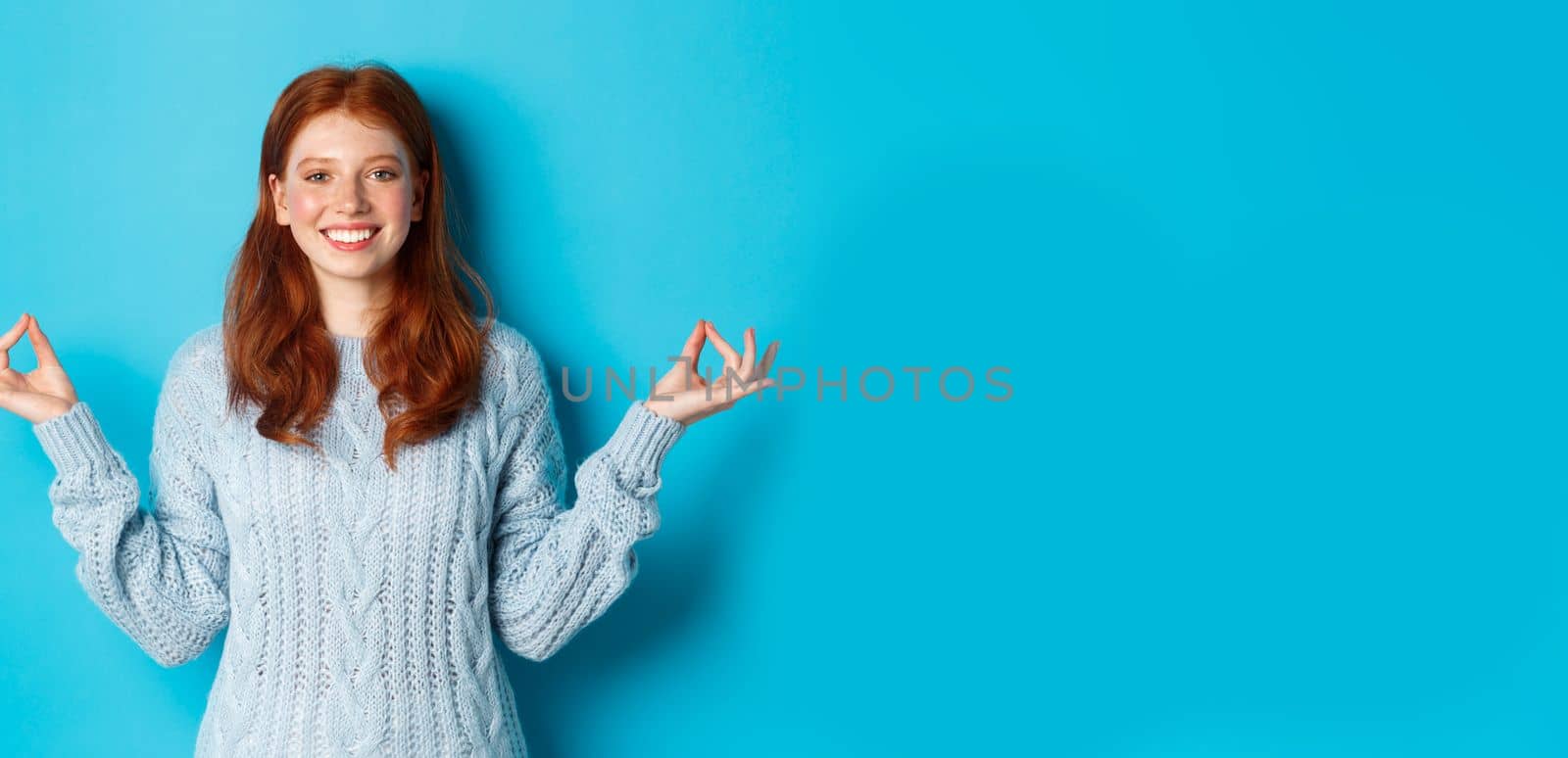 Smiling confident girl with red hair staying patient, holding hands in zen, meditation pose and staring at camera, practice yoga, standing calm against blue background.