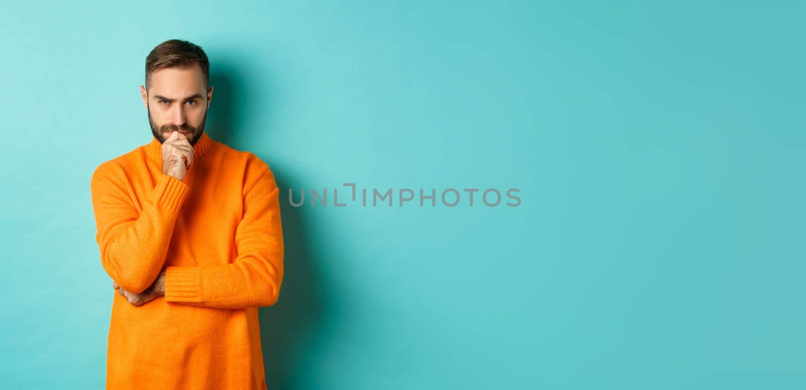 Thougtful young man making decision, looking serious and thinking, choosing, standing near copy space turquoise background.