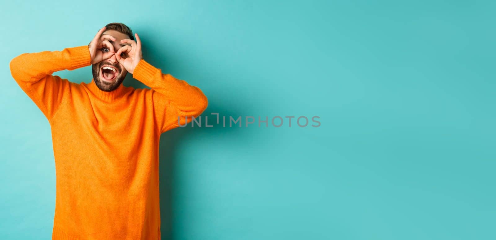 Image of funny and happy guy holding hands on eyes, looking through fake glasses, smiling amused, standing in orange sweater against turquoise background.