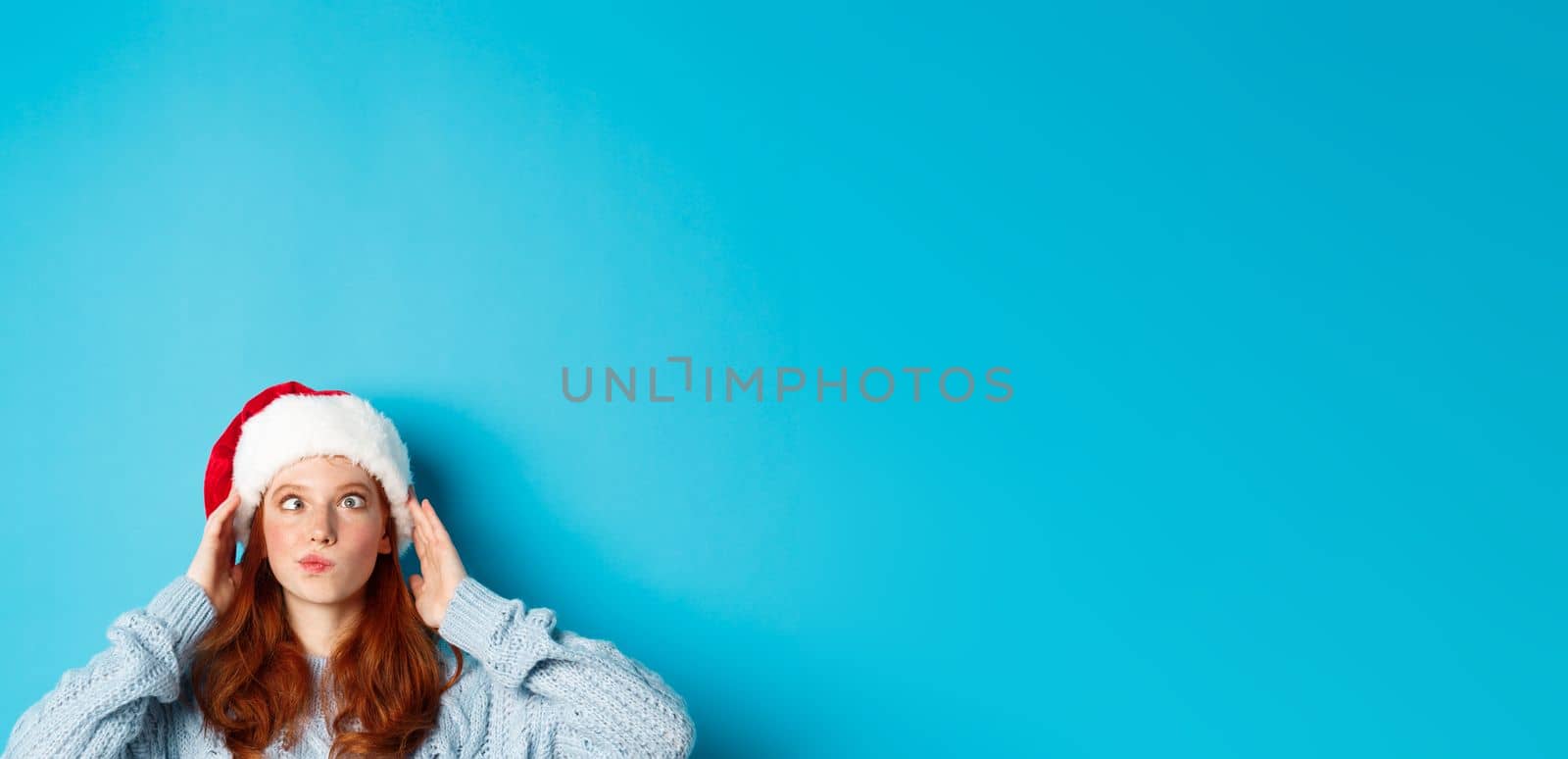 Winter holidays and Christmas eve concept. Head of funny redhead girl in santa hat, appear from bottom and squinting, making silly faces, standing near copy space on blue background.