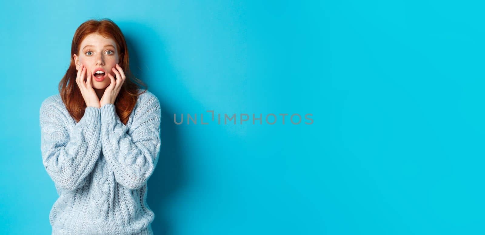 Startled redhead girl staring at something amazing, gasping in awe, standing in sweater against blue background. Copy space