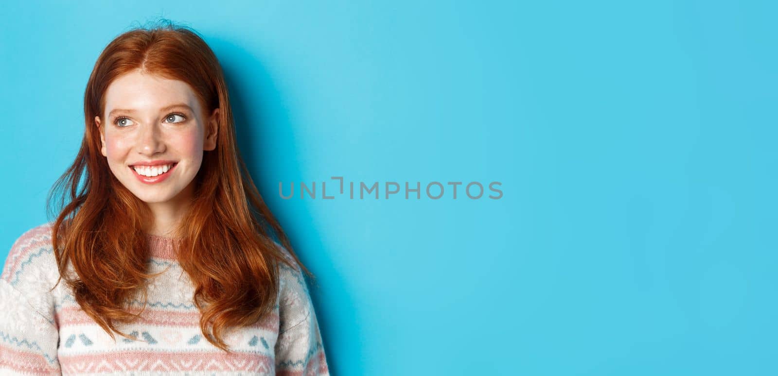 Close-up of beautiful redhead female model in winter sweater, smiling and looking left at copy space, standing over blue background by Benzoix